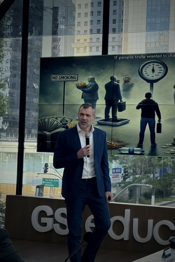 Roberto Monfort no palco de um evento na GS Educação, de terno, microfone na mão, com o telão do MindEquity ao fundo