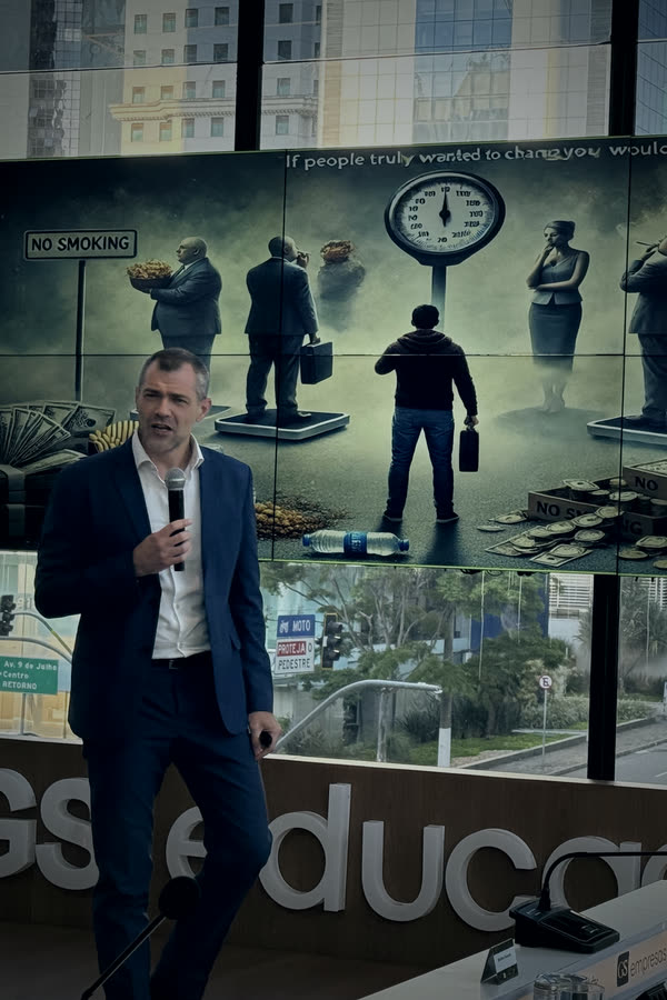 Roberto Monfort palestrando no palco de um evento na GS Educação, microfone na mão, telão ao fundo